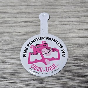 Puede incluir: Un botón redondo blanco con el personaje de dibujos animados Pink Panther en rosa, flexionando sus músculos. El botón dice "PINK PANTHER PAINLESS PIN" y "clean n treat MEDICATED FIRST AID PADS". Tiene una pestaña blanca.