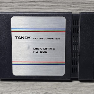 May include: A black Tandy Color Computer disk drive, model FD-500. The drive has a silver label with the Tandy logo and text, and is accented with blue, green, and red stripes. The drive has a rectangular shape with a ribbed side.