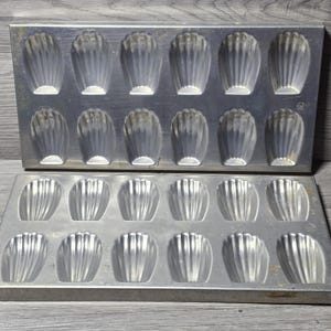 May include: Two rectangular silver-colored metal baking pans, each featuring twelve shell-shaped indentations arranged in two rows of six. The pans have a smooth, metallic finish with a distinct fluted pattern inside each individual mold cavity.