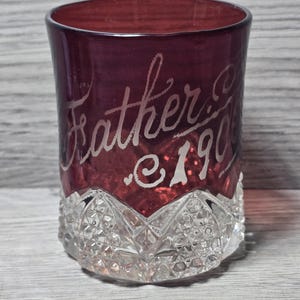 Könnte beinhalten: Ein tiefrotes Glas mit einem klaren, facettierten Boden im Diamantschliffmuster. Der obere Teil des Glases ist glatt und mit dem in Schreibschrift geätzten Text "Father" sowie der Jahreszahl "1906" in einer hellen, mattierten Optik versehen.