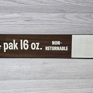 May include: A rectangular wooden sign with the text "4 pak 16 oz. NON-RETURNABLE" in white lettering. The sign has a dark brown finish and a white label area on the right side. Two small holes are visible near the top of the sign.
