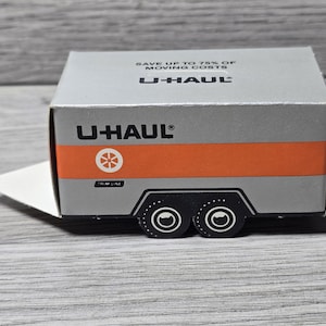 May include: A small, silver U-Haul box with an orange stripe and black trailer wheels. The U-Haul logo and text are visible on the box. The box has a white flap extending from the side. The text on the box says "Save up to 75% of moving costs."