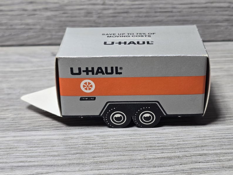 May include: A small, silver U-Haul box with an orange stripe and black trailer wheels. The U-Haul logo and text are visible on the box. The box has a white flap extending from the side. The text on the box says "Save up to 75% of moving costs."