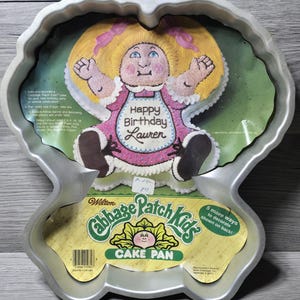 Puede incluir: Un molde para pastel vintage de Wilton Cabbage Patch Kids. El molde es de color plateado y tiene la forma de una muñeca Cabbage Patch Kid. La imagen en el molde muestra una muñeca con cabello rubio, un vestido rosa y el texto "Happy Birthday Lauren".