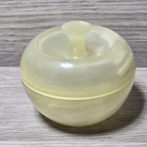 May include: A pale yellow, spherical container with a lid and a small handle on top. The container has a pearlescent, marbled appearance. The lid fits snugly on the base, creating a smooth, rounded shape. The container is sitting on a gray wooden surface.
