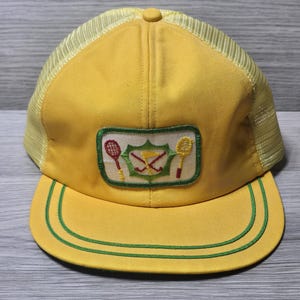 May include: A yellow trucker hat featuring a mesh back and a rectangular patch on the front. The patch displays a green shield emblem with crossed golf clubs and a flag, flanked by a red tennis racket on the left and a yellow racket on the right.