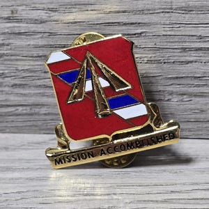 May include: A gold-colored pin with a red shield design, featuring white, blue, and gold accents. The pin has the words "MISSION ACCOMPLISHED" in gold lettering. The pin is likely a military or commemorative item.