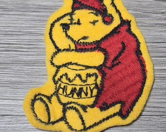 May include: Yellow felt Winnie the Pooh applique with black embroidery. Pooh is wearing a red felt hat and a red felt robe. He is holding a yellow felt honey pot with the word "HUNNY" embroidered in black.