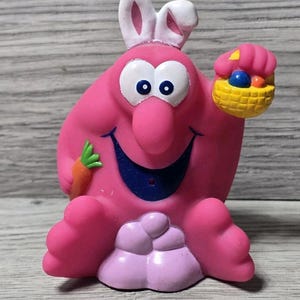 Puede incluir: Una figura rosa de Pascua con orejas de conejo, sosteniendo una zanahoria y una cesta de huevos coloridos. La figura tiene ojos grandes, una nariz azul y una cara sonriente. La figura está sobre una superficie de madera.