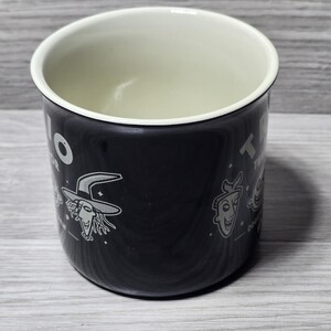 Puede incluir: Taza de esmalte negro con interior blanco. La taza presenta ilustraciones de personajes y texto. La taza est&aacute; sobre una superficie de madera gris claro.