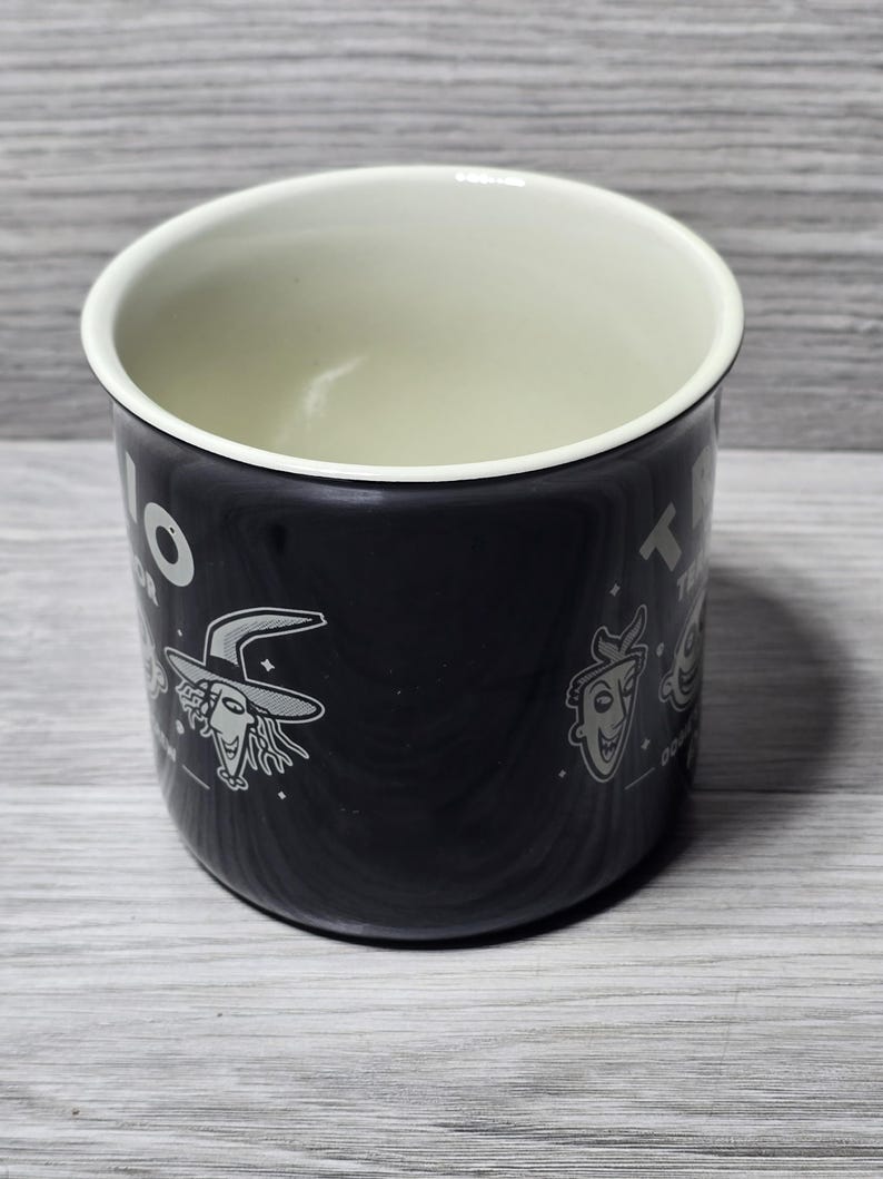 Puede incluir: Taza de esmalte negro con interior blanco. La taza presenta ilustraciones de personajes y texto. La taza est&aacute; sobre una superficie de madera gris claro.