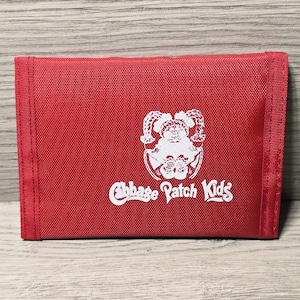Puede incluir: Una cartera roja de Cabbage Patch Kids con un logotipo blanco que presenta una ilustración de muñeca. La cartera es rectangular y plegada, con una superficie texturizada. El logotipo y el texto son blancos.
