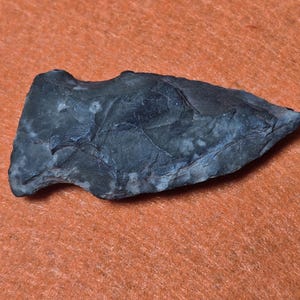 Puede incluir: Una punta de flecha gris oscuro con una superficie texturizada, probablemente hecha de piedra. La punta de flecha está sobre un fondo naranja. La punta de flecha tiene forma triangular con base dentada.