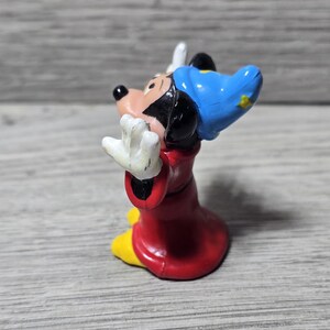 May include: A small figurine of Mickey Mouse in a sorcerer's outfit. The figure is wearing a red robe, yellow shoes, a blue hat with a yellow star, and white gloves. The figure is posed with arms outstretched.
