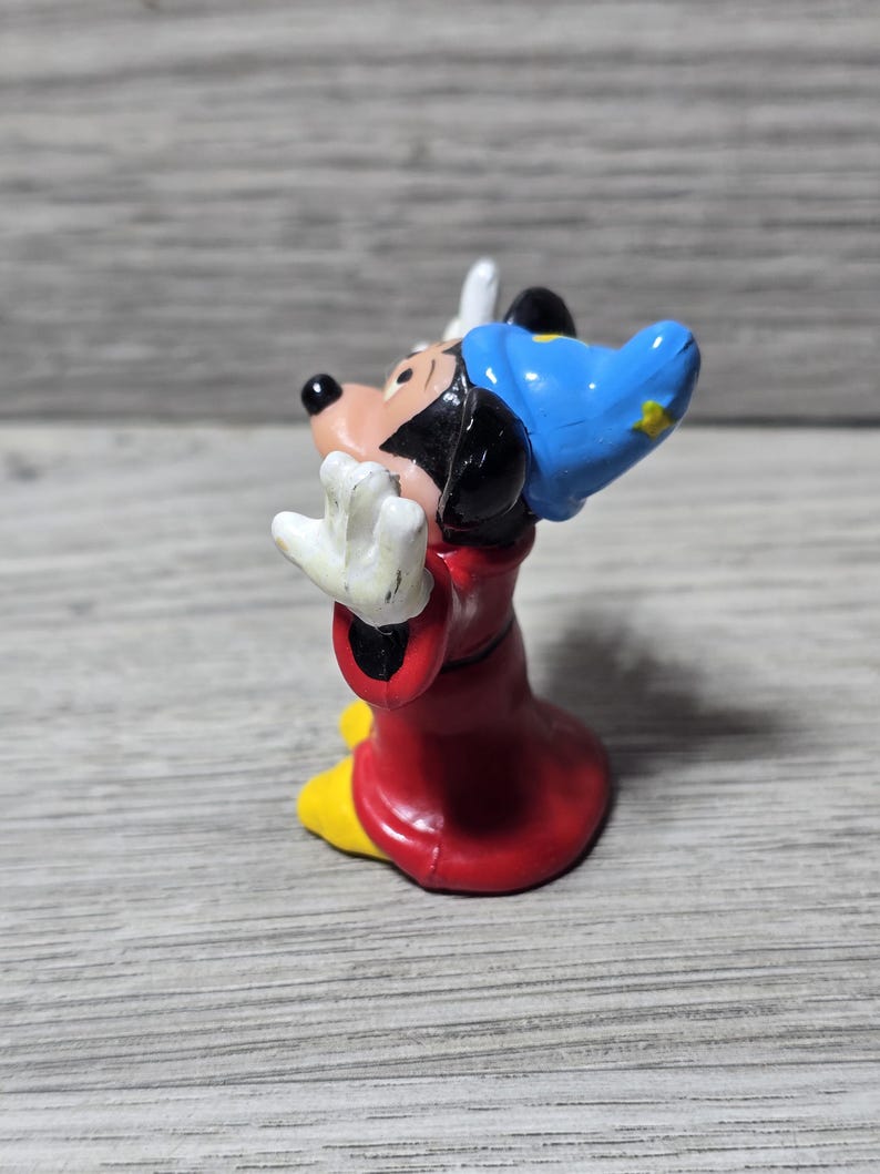 May include: A small figurine of Mickey Mouse in a sorcerer's outfit. The figure is wearing a red robe, yellow shoes, a blue hat with a yellow star, and white gloves. The figure is posed with arms outstretched.