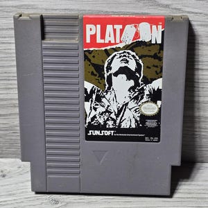 以下が含まれることがあります： 「Platoon」というゲームの、グレーの任天堂エンターテインメントシステム（NES）カートリッジ。ラベルには、上を見上げる男性の白黒画像があり、タイトルは赤と白で表示されています。Sunsoftのロゴも表示されています。