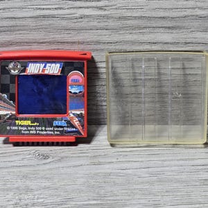 May include: A red and black Tiger Electronics Indy 500 handheld game with a clear plastic case. The game features a blue screen and the text "INDY-500" and "TIGER." The game is from 1996 and licensed by IMS Properties, Inc.