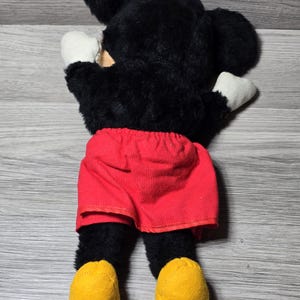 May include: A black plush Mickey Mouse toy with red shorts and yellow shoes. The toy is lying on its back with its arms outstretched.