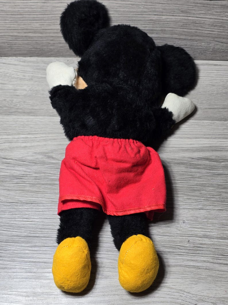 May include: A black plush Mickey Mouse toy with red shorts and yellow shoes. The toy is lying on its back with its arms outstretched.