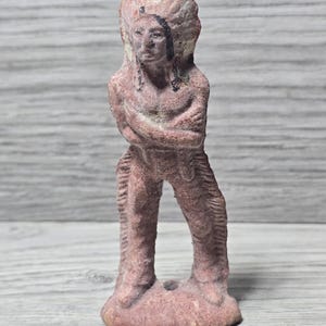 May include: A small, weathered statue of a Native American chief in a headdress. The figure is a muted reddish-brown color, with arms crossed and detailed clothing. The statue is standing on a flat base.