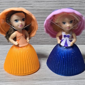 Puede incluir: Dos pequeñas figuras de muñecas. Una muñeca viste un vestido y sombrero naranja, la otra un vestido azul y sombrero rosa. Ambas muñecas tienen ojos azules y llevan una mascarilla. Las muñecas están sobre una superficie gris claro.