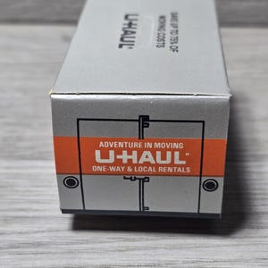 May include: A grey U-Haul box with orange and white text reading "Adventure in Moving" and "U-Haul One-Way & Local Rentals". The box also displays the U-Haul logo and the text "Save up to 75% of Moving Costs".
