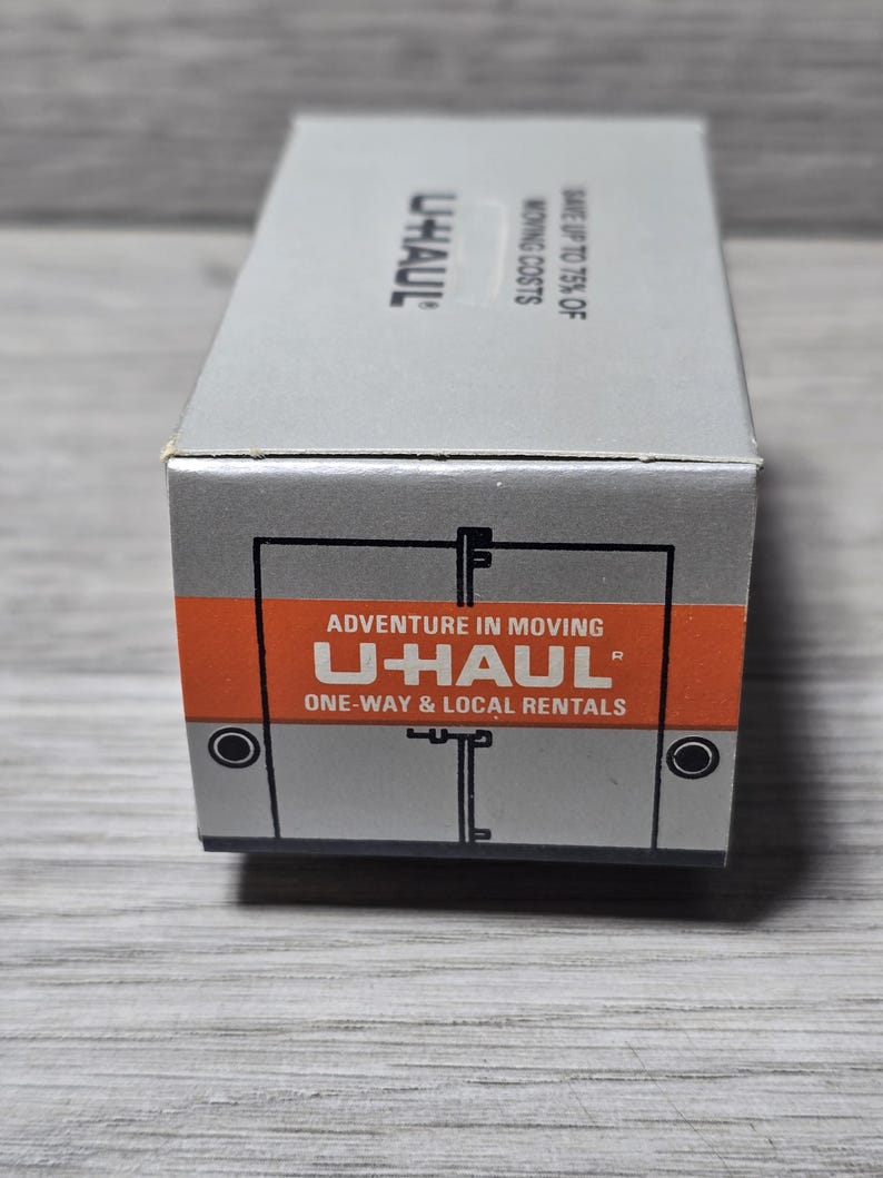 May include: A grey U-Haul box with orange and white text reading "Adventure in Moving" and "U-Haul One-Way & Local Rentals". The box also displays the U-Haul logo and the text "Save up to 75% of Moving Costs".