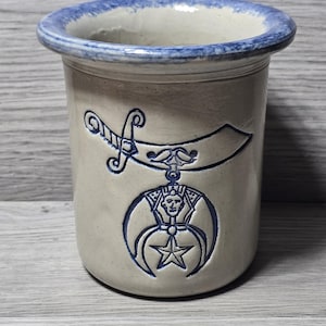 Puede incluir: Un jarrón de cerámica con cuerpo beige claro y borde azul. El jarrón presenta un emblema azul con una espada, una luna creciente, una estrella y una cara humana. El jarrón es cilíndrico y se encuentra sobre una superficie de madera.