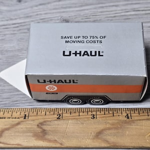 May include: A miniature U-Haul trailer model with a silver and orange design. The box reads "SAVE UP TO 75% OF MOVING COSTS" and features the U-Haul logo. A ruler in inches is in the foreground.