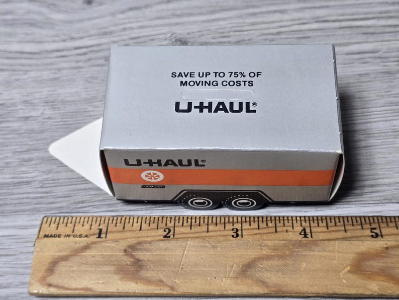 May include: A miniature U-Haul trailer model with a silver and orange design. The box reads "SAVE UP TO 75% OF MOVING COSTS" and features the U-Haul logo. A ruler in inches is in the foreground.