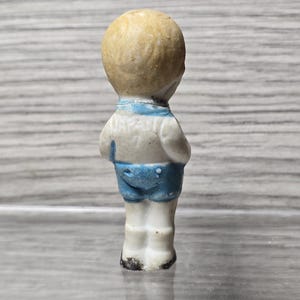 Puede incluir: Peque&ntilde;a figura vintage de un ni&ntilde;o, vista desde atr&aacute;s. La figura tiene una cabeza de color marr&oacute;n claro y lleva una camisa blanca con ribete azul y pantalones cortos azules. La palabra "JAPAN" es visible en la camisa.