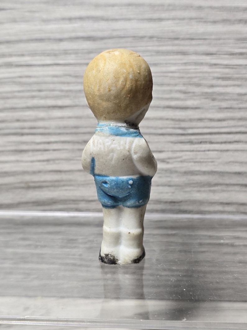 Puede incluir: Peque&ntilde;a figura vintage de un ni&ntilde;o, vista desde atr&aacute;s. La figura tiene una cabeza de color marr&oacute;n claro y lleva una camisa blanca con ribete azul y pantalones cortos azules. La palabra "JAPAN" es visible en la camisa.