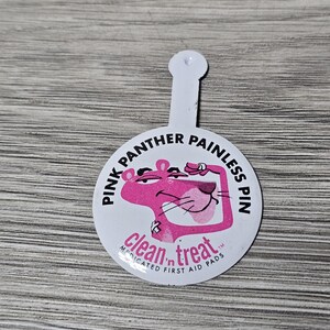 Può includere: Una spilla Pink Panther Painless bianca e rosa. La spilla presenta una pantera rosa dei cartoni animati con la scritta "Pink Panther Painless Pin" e "clean 'n treat medicated first aid pads". La spilla ha un manico bianco.