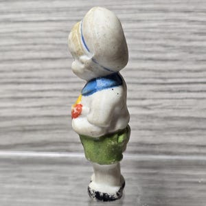 Pu&ograve; includere: Piccola statuina vintage in porcellana di un bambino. La statuina indossa un completo da marinaio bianco con dettagli blu, una gonna verde e un cappello bianco con una striscia blu. La statuina tiene in mano un oggetto rosso.
