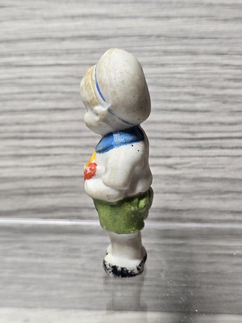 Pu&ograve; includere: Piccola statuina vintage in porcellana di un bambino. La statuina indossa un completo da marinaio bianco con dettagli blu, una gonna verde e un cappello bianco con una striscia blu. La statuina tiene in mano un oggetto rosso.