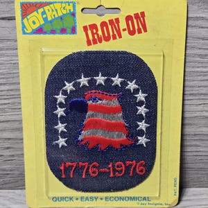 May include: An iron-on patch featuring a patriotic design. The patch has a blue background with white stars and a red, white, and blue emblem. The text "1776-1976" is displayed below the emblem. The patch is packaged on a yellow backing.