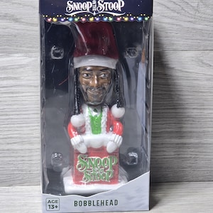 May include: A bobblehead figure of a man dressed in a Santa Claus outfit. The figure has a red hat and coat with white trim, and a green vest. The base of the figure has the words "Snoop on the Stoop" and "Bobblehead" printed on it.