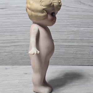 May include: A vintage bisque doll figurine, featuring a nude child with a detailed face and curly blonde hair. The doll has rosy cheeks and is standing upright. The figurine is a collectible item.