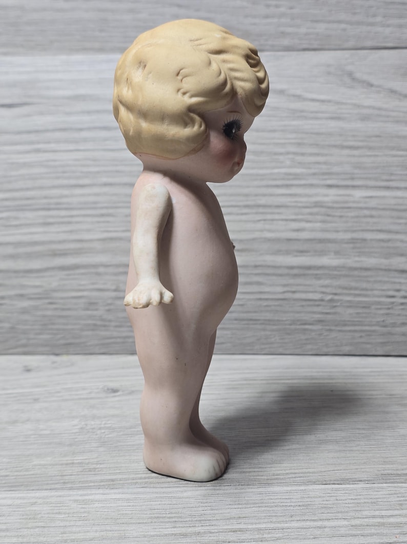 May include: A vintage bisque doll figurine, featuring a nude child with a detailed face and curly blonde hair. The doll has rosy cheeks and is standing upright. The figurine is a collectible item.
