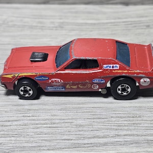 May include: A red die-cast toy car featuring a black hood scoop, blue tinted windows, and various racing decals including Goodyear, Champion, and Union logos. The vehicle has silver-colored wheels and shows signs of paint wear along the edges.