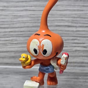 Puede incluir: Figura de dibujos animados naranja con ojos blancos grandes, sosteniendo un patito de goma amarillo y un cepillo de dientes. La figura lleva pantalones cortos azules y está de pie sobre un bloque rectangular blanco.