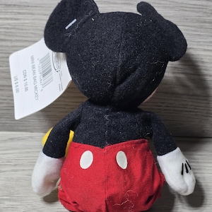 以下が含まれることがあります： 黒い耳、黒い体、赤いショートパンツ、白い手袋が付いた小さなミッキーマウスのぬいぐるみ。おもちゃには「MINI BEAN BAG MICKEY」と書かれたタグが付いています。ぬいぐるみは木製の表面にあります。