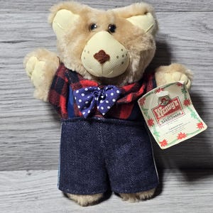 Op de afbeelding: Een pluchen teddybeer met een rood en zwart geruit overhemd, een blauwe strik met witte stippen en een spijkerbroek. De beer heeft een lichtbruine snuit en oren. Een Wendy's-label is bevestigd.