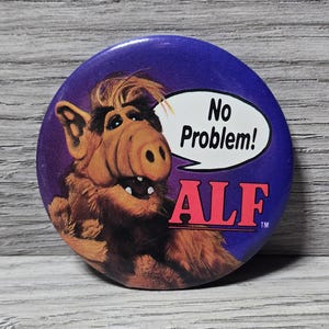 May include: A round, purple button featuring the character ALF. The image shows ALF with a speech bubble that says "No Problem!" and the name "ALF" in red letters.
