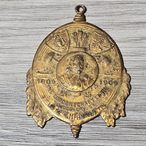May include: A gold-coloured metal medallion featuring a central portrait and the text "The Tercentenary Celebration" and "Hampton". The dates 1609 and 1909 are also visible. The medallion has a loop at the top for hanging.