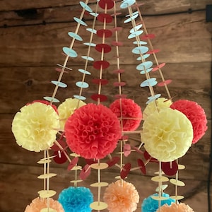 May include: A colorful hanging mobile featuring paper pom-poms in shades of yellow, red, blue, and orange. The mobile is suspended from a series of wooden dowels and decorative paper circles, creating a festive and eye-catching design.