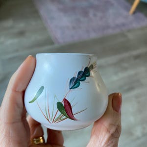 May include: A small, white ceramic container with a rounded shape. It features a hand-painted design of green, teal, and red leaf-like shapes. The container is held in a person's hand.