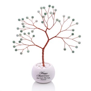 Puede incluir: Una escultura de árbol decorativa con ramas de alambre de cobre trenzado adornadas con pequeñas cuentas de piedra redondas de color verde pálido. El árbol está en una maceta de cerámica blanca esférica llena de piedras blancas, con la inscripción "Happy Anniversary, One Love, One Life, Forever."