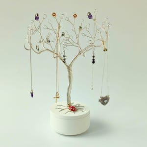 May include: A silver-toned jewellery tree with a tree trunk design, displaying necklaces and earrings. The tree features small decorative elements in purple, green, and gold. A white, round base with a ladybug design supports the tree.