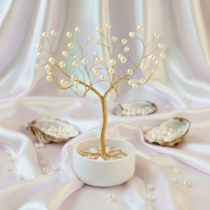 Puede incluir: Una escultura decorativa de árbol con tronco y ramas doradas adornadas con perlas blancas. El árbol está en una maceta de cerámica blanca llena de pequeñas piedras blancas. Conchas de ostra llenas de perlas están dispersas.